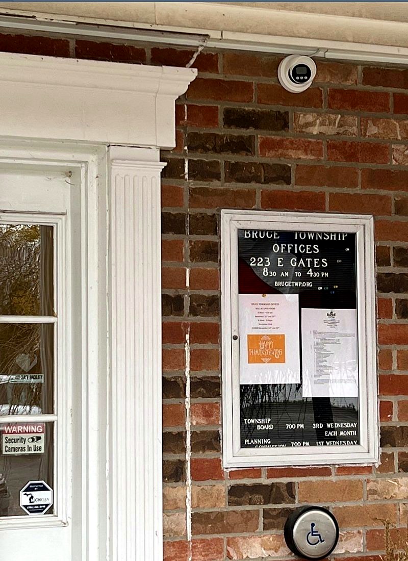 Exterior dome security camera mounted on the brick wall above the entrance at Bruce Township offices, 223 E Gates in Romeo, Michigan, with Michigan Security Systems camera-in-use decals, installed by Michigan Security Systems