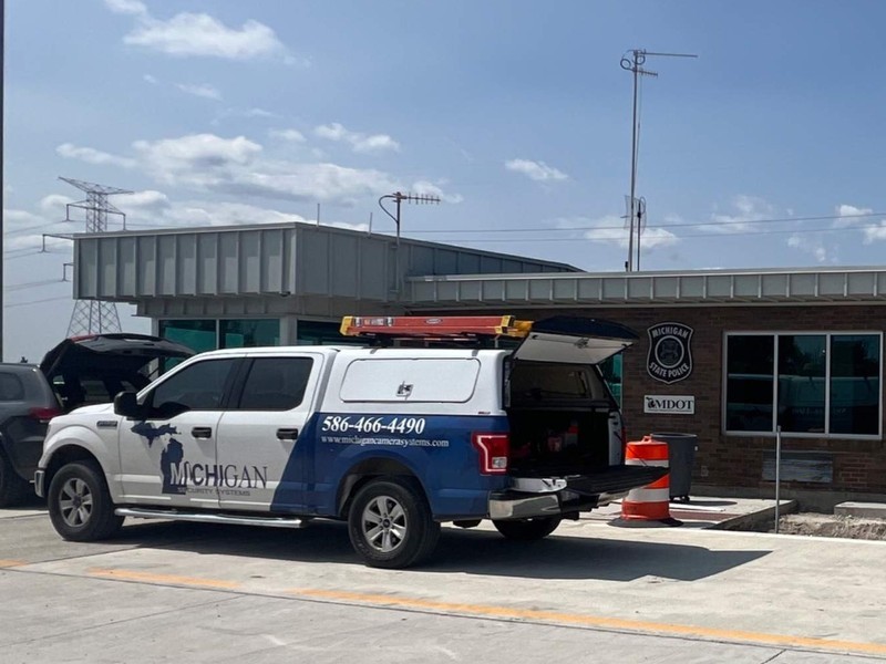 Michigan Security Systems service truck on site at a commercial security camera installation, with a wall-mounted dome camera under the building roofline
