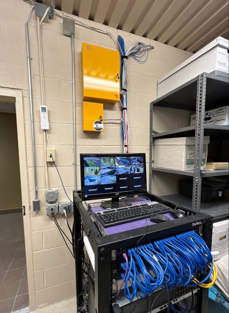 PDK access control panels, live security camera monitor, and structured network cabling rack inside the equipment room at Bruce Township City Hall in Romeo, Michigan, installed by Michigan Security Systems