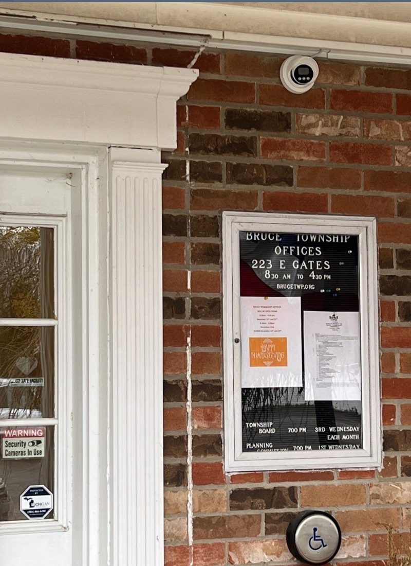 Exterior dome security camera mounted on the brick wall above the entrance at Bruce Township offices, 223 E Gates in Romeo, Michigan, with Michigan Security Systems camera-in-use decals, installed by Michigan Security Systems