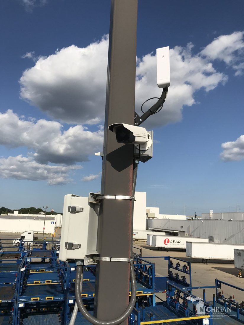 Pole-mounted outdoor bullet security camera with a Ubiquiti wireless network bridge and weatherproof equipment enclosure overlooking the equipment yard at Merlo Construction in Milford, Michigan, installed by Michigan Security Systems