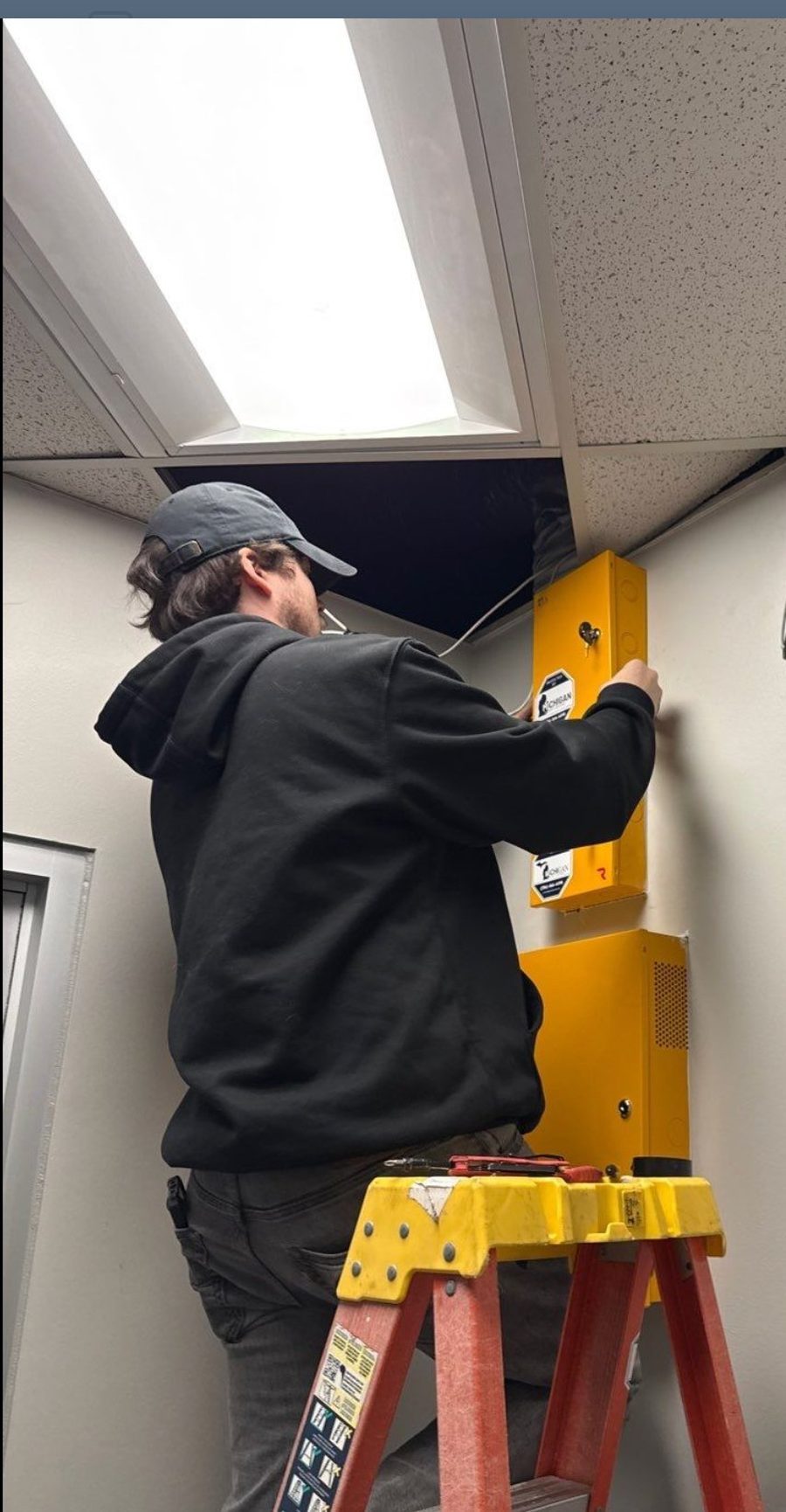 Michigan Security Systems technician on a ladder wiring yellow security enclosures with Protected By decals at the J.G. Kern Enterprises automotive plant in Sterling Heights, Michigan