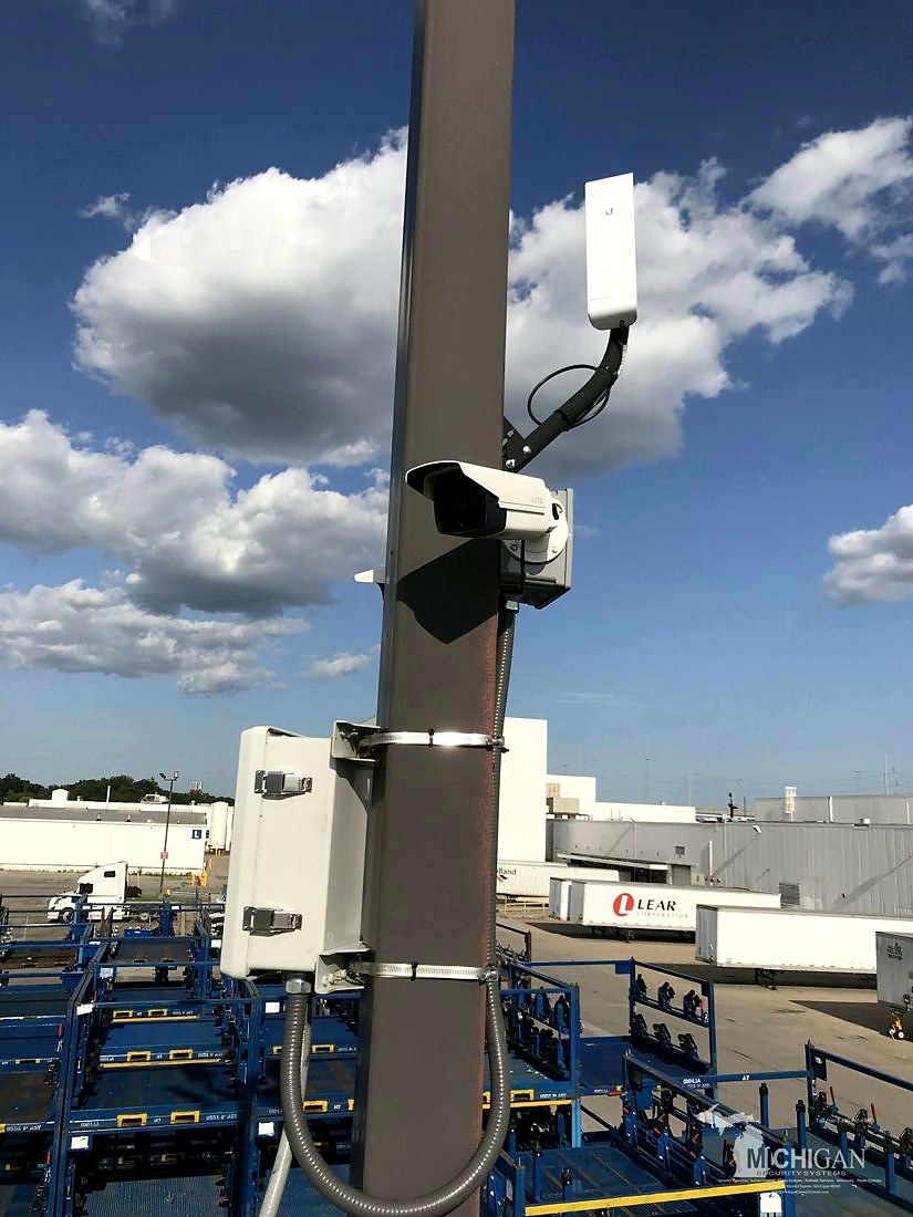 Pole-mounted outdoor bullet security camera with a Ubiquiti wireless network bridge and weatherproof equipment enclosure overlooking the equipment yard at Merlo Construction in Milford, Michigan, installed by Michigan Security Systems